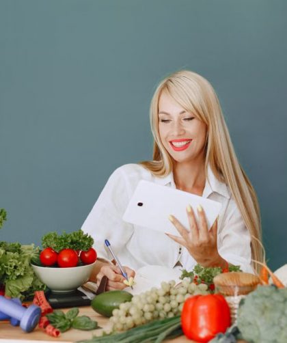 Woman using a tablet surrounded by fresh vegetables at a table, showcasing healthy lifestyle and technology.