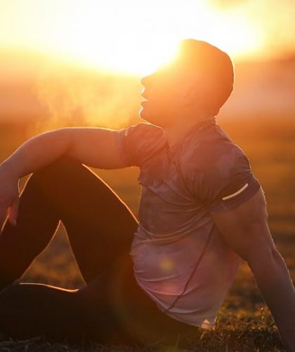 Muscular man sits in rural field at sunset, exhaling visible breath.