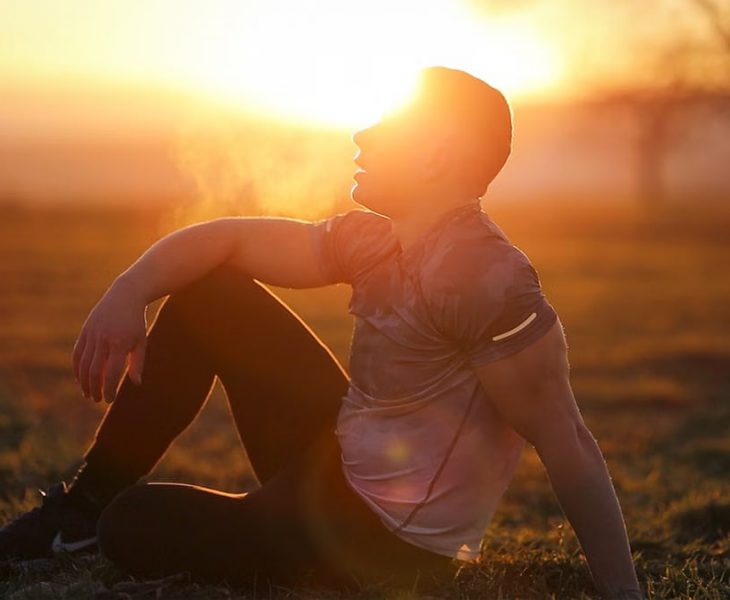 Muscular man sits in rural field at sunset, exhaling visible breath.
